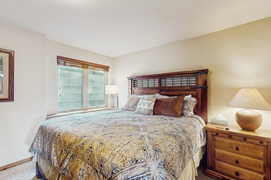 A bedroom with a large bed, a nightstand, a lamp, and a window with blinds.