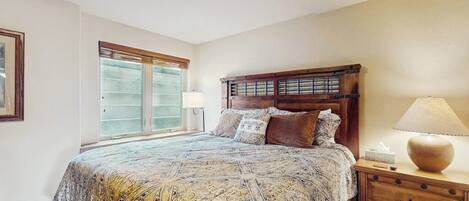 A bedroom with a large bed, a nightstand, a lamp, and a window with blinds.