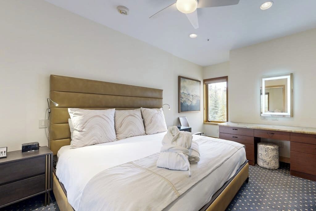 A hotel room with a bed, a bedside table, a desk with a mirror, a ceiling fan, and a window.