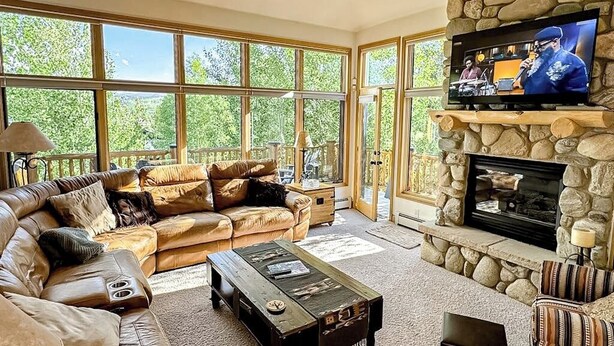 A living room with a stone fireplace, a television, and a large window overlooking greenery. - Enchanting mountain oasis with hot tub, fireplaces, deck, & views (Fraser)