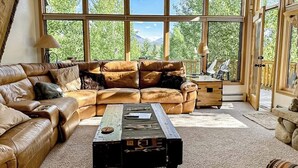 A spacious living room with a large window, leather sofa, wooden coffee table, and a view of greenery outside. - Enchanting mountain oasis with hot tub, fireplaces, deck, & views (Fraser)