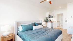 Un dormitorio moderno con una cama grande, una mesita de noche, un ventilador de techo y vistas a la cocina contigua.