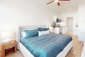 A modern bedroom with a large bed, a nightstand, a ceiling fan, and a view into the adjacent kitchen. - Modern ground-floor studio with mountain view, kitchenette & parking (Fraser)