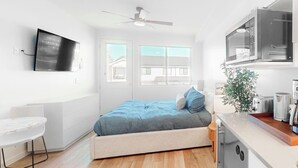 Una habitación pequeña y funcional con cama, un escritorio, televisor y kitchenette.
