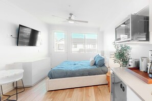 A compact bedroom with a bed, a small desk, a TV, and a kitchenette. - Modern ground-floor studio with mountain view, kitchenette & parking (Fraser)