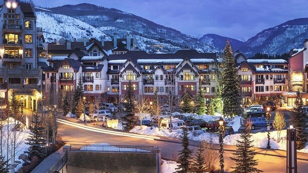 A resort with multiple-story buildings, a parking area, and snow-covered mountains in the background. - Ski-in/ski-out condo w/ balcony, fireplace, fast WiFi, W/D & shared pool/hot tub (Vail)