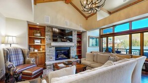 A spacious living room with a stone fireplace, a large sofa, a plush armchair, and a dining area with large windows. - Ski-in/ski-out condo w/ balcony, fireplace, fast WiFi, W/D & shared pool/hot tub (Vail)