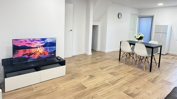 Ett modernt vardagsrum med en platt-TV, en matplats med bord och stolar samt ett köksområde med kylskåp.