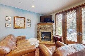 A living room with leather sofas, a stone fireplace, a flat-screen TV, and a large window with blinds. - Antlers Gulch Townhouse Next to National Forest & Golf w/shared Hot Tub, WiFi (Keystone)
