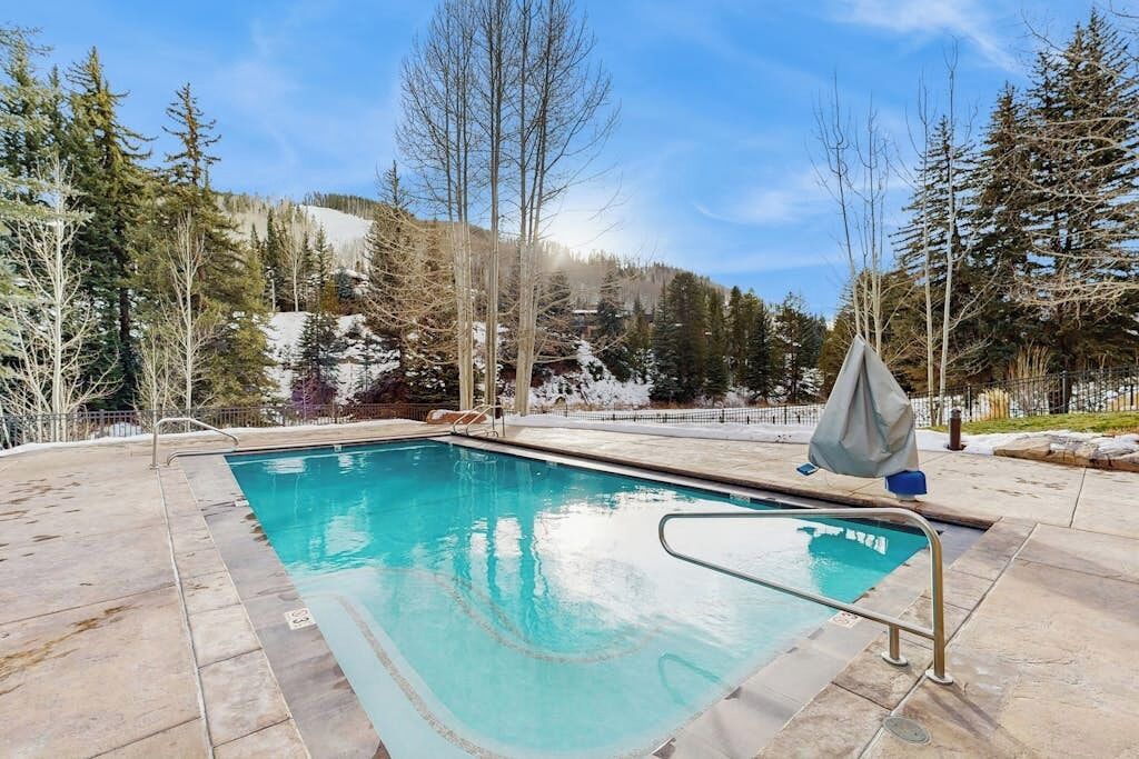 A swimming pool with clear blue water, surrounded by a stone deck and trees.