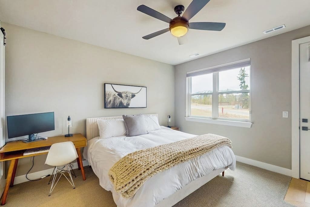 A bedroom with a bed, a desk with a computer, a chair, a ceiling fan, a window, and a picture on the wall.