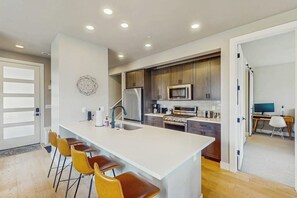 A modern kitchen with a central island, bar stools, and a dining area with a computer and chair. - Inviting Condo with Mountain Views, Fireplace & Space for the Entire Family (Fraser)