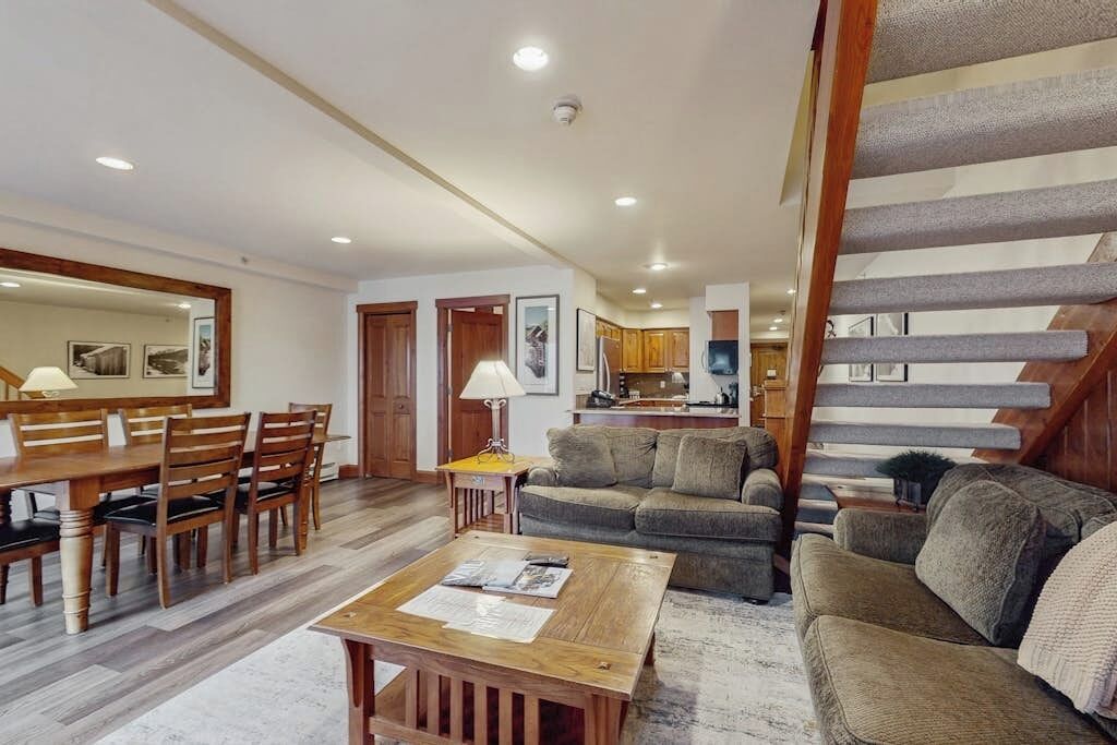 A spacious living area with a dining table, multiple sofas, a staircase, and a kitchen in the background.