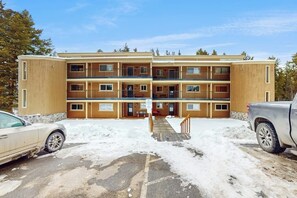 A multi-story building with a central entrance, a parking area with snow, and a truck parked to the right. - Beaver Village condo with heated bathroom floors and free ski shuttle (Winter Park)