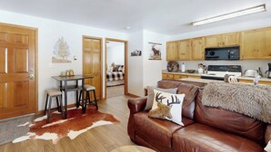 A cozy living room with a brown leather couch, a dining table with stools, and a kitchen area with wooden cabinets and a microwave.