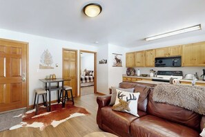 A cozy living room with a brown leather couch, a dining table with stools, and a kitchen area with wooden cabinets and a microwave.