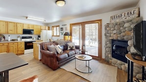 A kitchen with wooden cabinets, a microwave, and a refrigerator. There is a brown leather sofa with pillows, a round coffee table, and a stone fireplace.