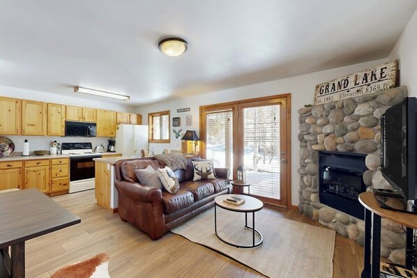 A kitchen with wooden cabinets, a microwave, and a refrigerator. There is a brown leather sofa with pillows, a round coffee table, and a stone fireplace.