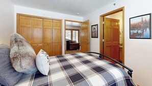 A bedroom with a bed, a wooden wardrobe, a door leading to another room, and a framed picture on the wall.