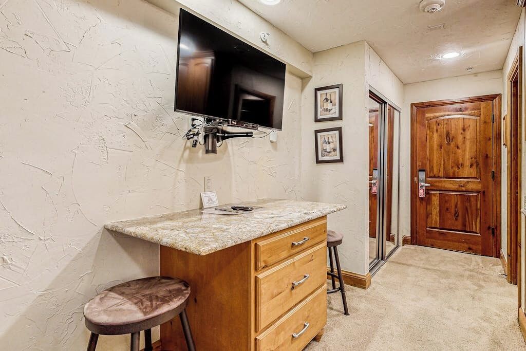 A bathroom with a granite countertop, wooden vanity, two stools, a mounted TV, and a wooden door.