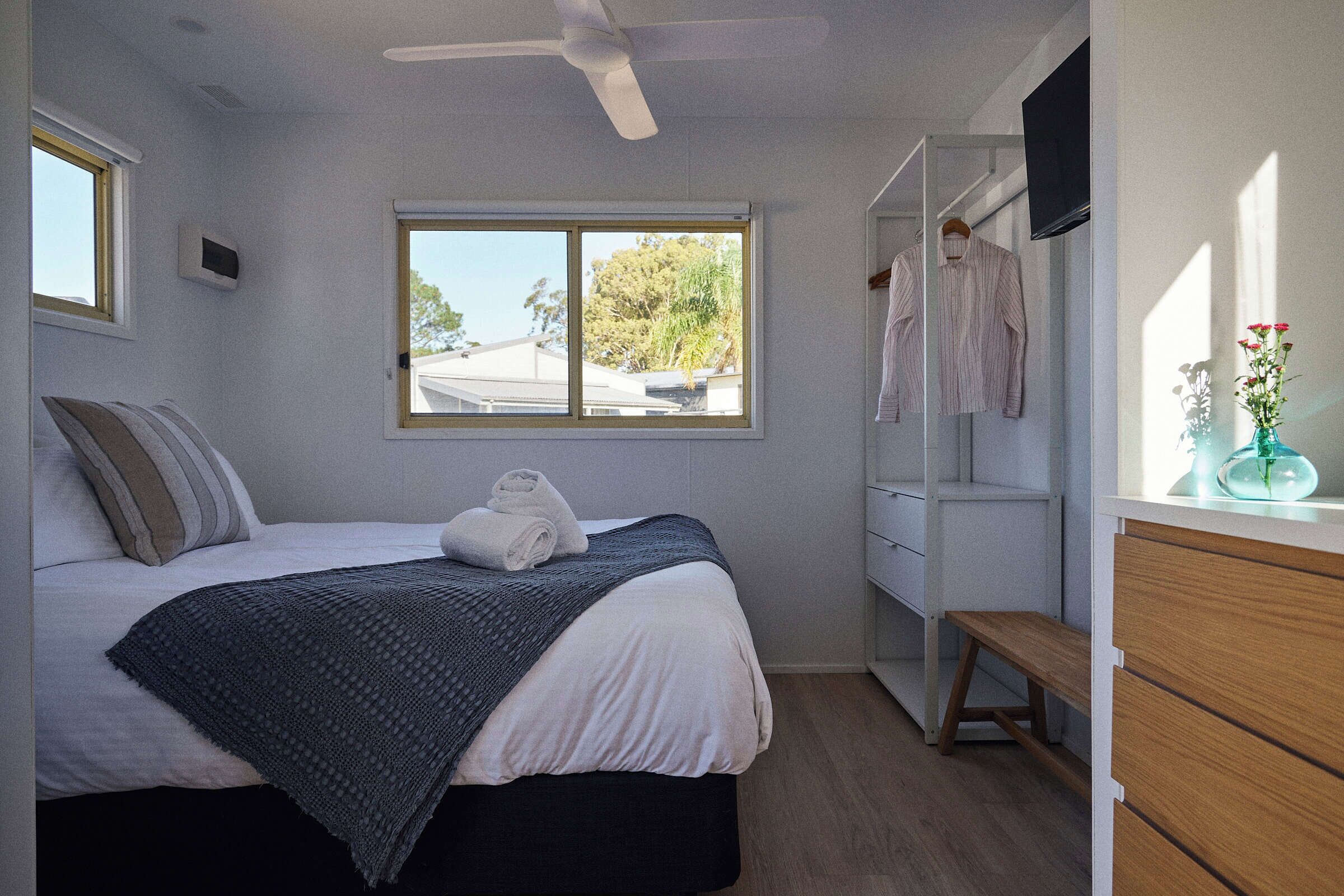 A bedroom with a bed, a window, a ceiling fan, a wardrobe, and a bench.