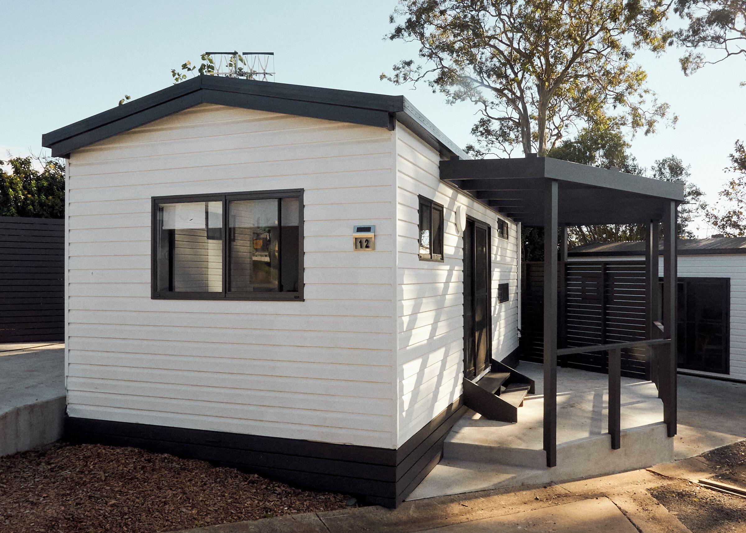 A small, single-story mobile home with a gabled roof, a front porch, and a number '12' sign.