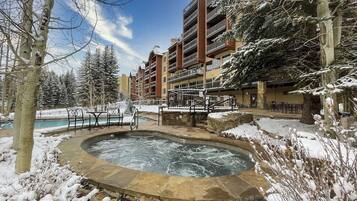 A hotel with a hot tub surrounded by snow, a pool, and a view of a mountain.