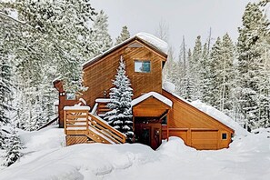Eine Holzblockhütte mit Treppe, einer kleinen Veranda und einem mit Schnee bedeckten Dach, umgeben von einem Wald.
