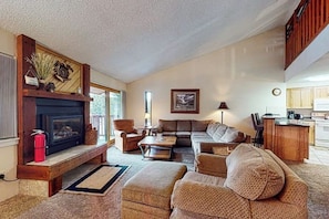 A cozy living room with a fireplace, a sofa set, and a kitchen area visible through an open door. - Gorgeous ski-in 3rd-floor condo with great views, hot tubs, fireplace, gym (Breckenridge)
