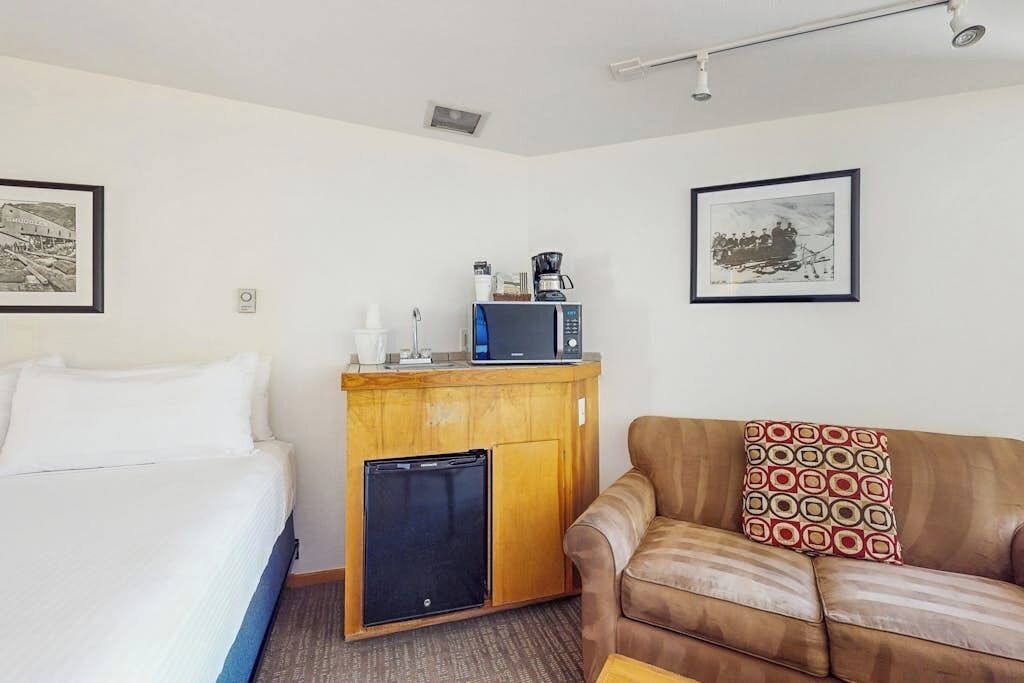Une chambre d’hôtel avec un lit, une petite kitchenette équipée d’un four à micro-ondes et d’un réfrigérateur, un canapé en cuir marron et deux tableaux encadrés accrochés aux murs.