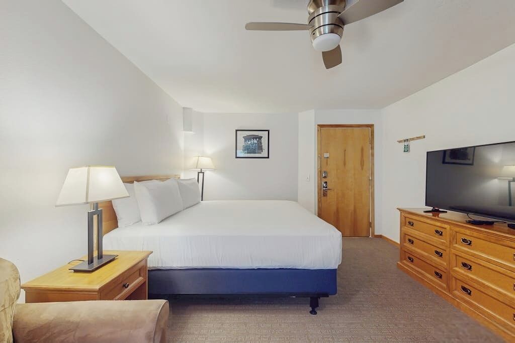 A hotel room with a bed, a television, a wooden dresser, a nightstand with a lamp, a ceiling fan, and a door.