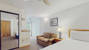 A hotel room with a bed, a chair, a desk with a lamp, a framed picture on the wall, and a ceiling fan.