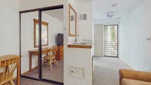A room with a desk, chair, and a mirror. There is a sliding door leading to another room with a bed and a window.