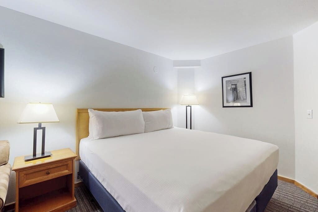 A hotel room with a bed, two bedside lamps, a nightstand, a framed picture on the wall, and a carpeted floor.