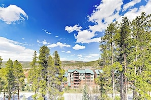 Ein mehrstöckiges Hotel mit Gründach, umgeben von hohen Kiefern, vor einer Bergkulisse und blauem Himmel.
