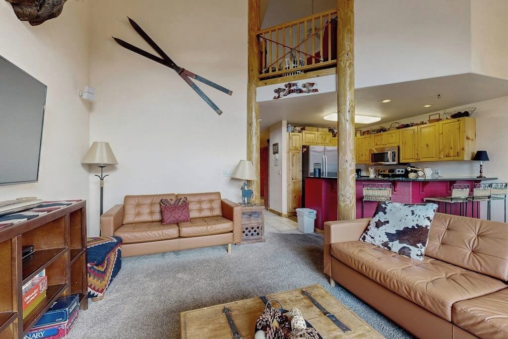 A living room with a sofa, coffee table, and bookshelf. There is a kitchen visible in the background.