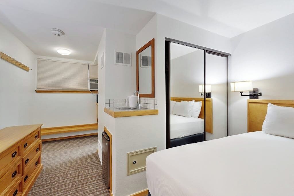 Una camera d'albergo con un letto, una cassettiera, uno specchio, una finestra con tende e una porta.