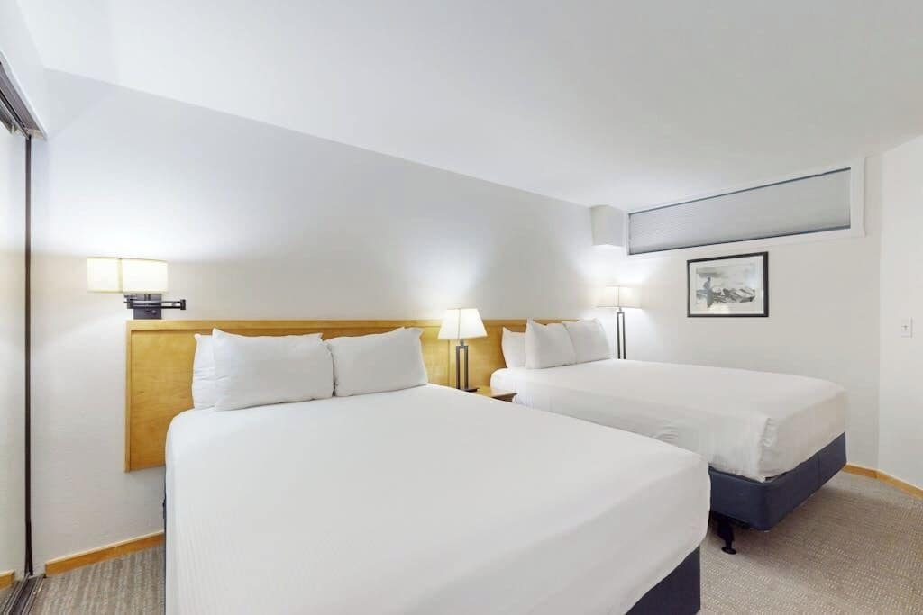 Una camera d'albergo con due letti, una testiera in legno, lampade sui comodini, un quadro appeso al muro e un pavimento con moquette.