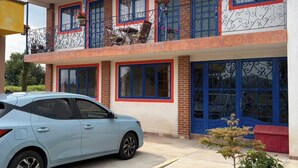 Un edificio de varios pisos con balcones, un coche estacionado adelante y un pequeño jardín.