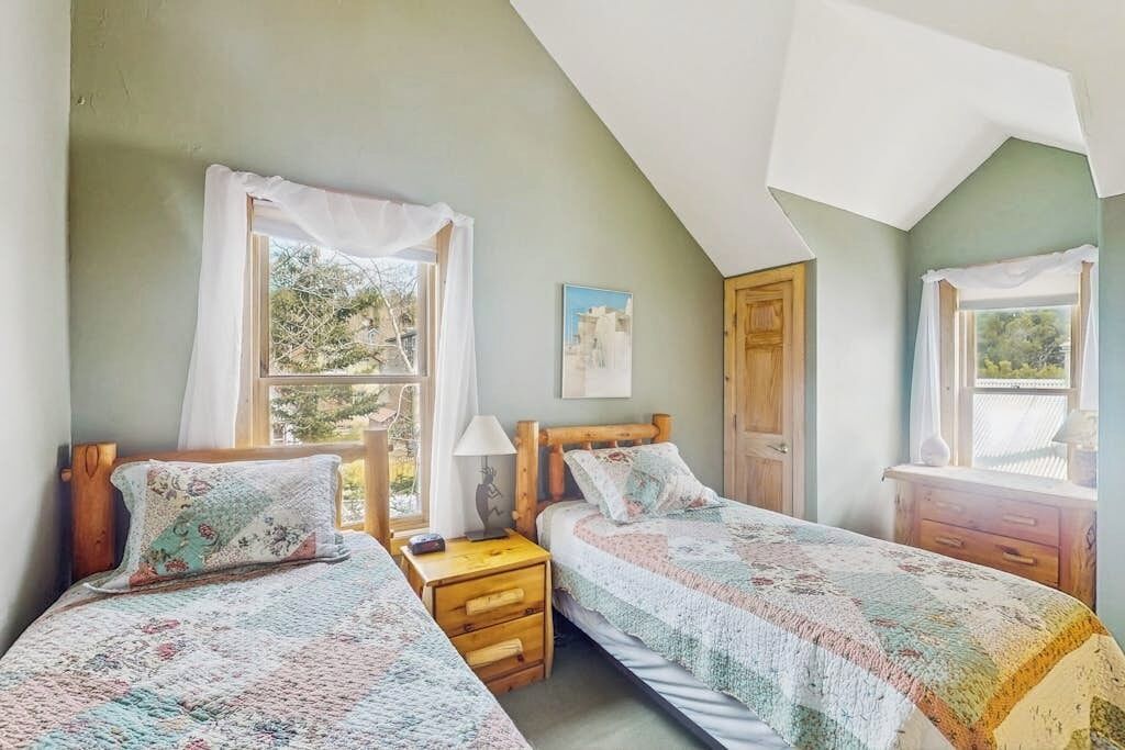 Una habitación con dos camas individuales, una mesita de noche de madera, una pequeña mesa y una ventana con cortinas.