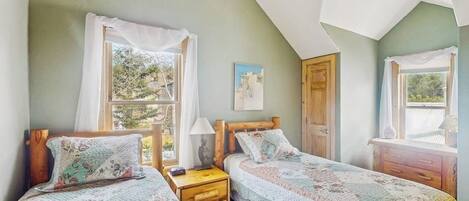 Una habitación con dos camas individuales, una mesita de noche de madera, una pequeña mesa y una ventana con cortinas.