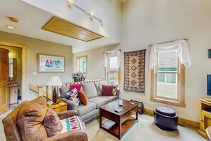 Ein Wohnzimmer mit Sofa, Couchtisch und einem kleinen Tisch mit Lampe. Es gibt ein Fenster mit Vorhängen, einen an der Wand montierten Fernseher und einen Teppich.