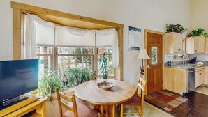Una habitación con televisión, una mesa redonda de madera, sillas, un ventanal con cortinas, una puerta y un área de cocina con armarios y un lavabo.