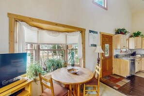 Ein Zimmer mit Fernseher, einem runden Holztisch, Stühlen, einem Fenster mit Vorhängen, einer Tür und einer Küchenzeile mit Schränken und Spüle.