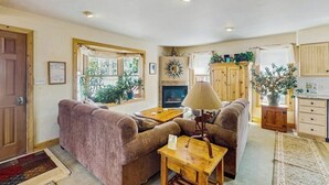 Una sala de estar acogedora con un sofá, una mesa y una lámpara. Hay una chimenea, una ventana con cortinas, y una zona de cocina con armarios y un fregadero.