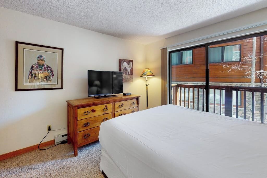 A hotel room with a bed, a TV on a wooden dresser, a lamp, and a large window with a view of a building.