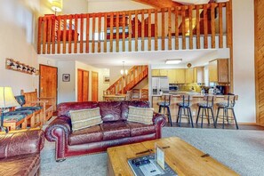 Ein großzügiger Wohnbereich mit hoher Holzbalkendecke, einem Ledersofa, einem Holztisch und einer Küche im Hintergrund.