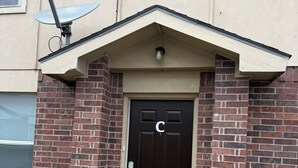 Un edificio de ladrillo con una puerta marrón oscuro marcada con una 'C', una antena parabólica y un pequeño escalón que lleva a la entrada.