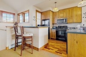 A kitchen with wooden cabinets, a microwave, a stove, and a bar area with chairs. - Ski-in/out condo with amazing views, washer/dryer, fireplace, & hot tub (Granby)