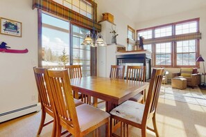 A dining area with a wooden table and chairs, a fireplace, and large windows with a view of the outdoors. - Ski-in/out condo with amazing views, washer/dryer, fireplace, & hot tub (Granby)